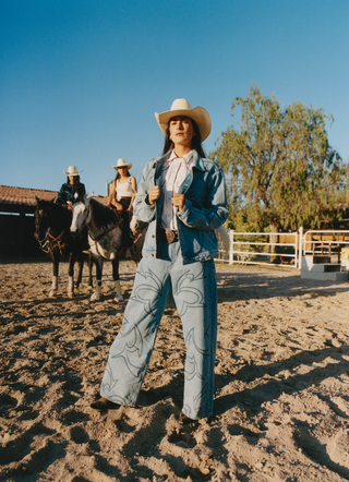 Mujer con chamarra de mezclilla y sombrero vaquero en un rancho, con un jinete a caballo al fondo